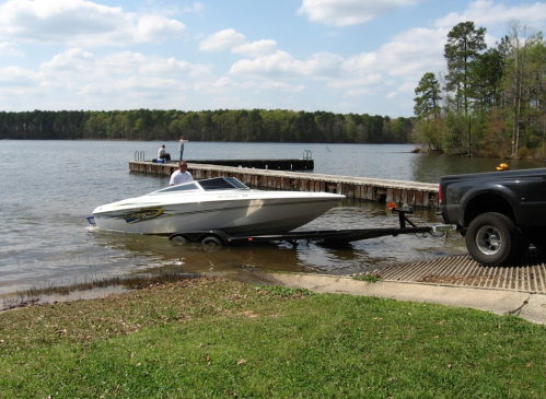 A white boat is being launched into a lake from a trailer, with a dock and trees in the background.