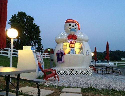 A whimsical snowman-shaped ice cream stand with lights, an "OPEN" sign, and outdoor seating in a rural setting.