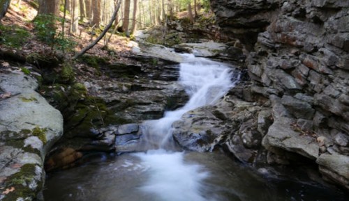 A serene waterfall cascades over rocky terrain, surrounded by lush greenery in a tranquil forest setting.