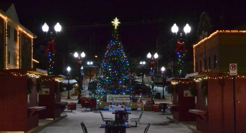 A brightly lit Christmas tree adorned with colorful lights stands in a festive plaza at night, surrounded by decorated buildings.