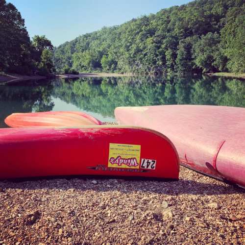 Two colorful kayaks resting on a gravel shore by a calm, reflective lake surrounded by lush green trees.