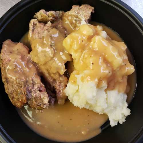 A black bowl containing meatloaf with gravy and a serving of mashed potatoes.