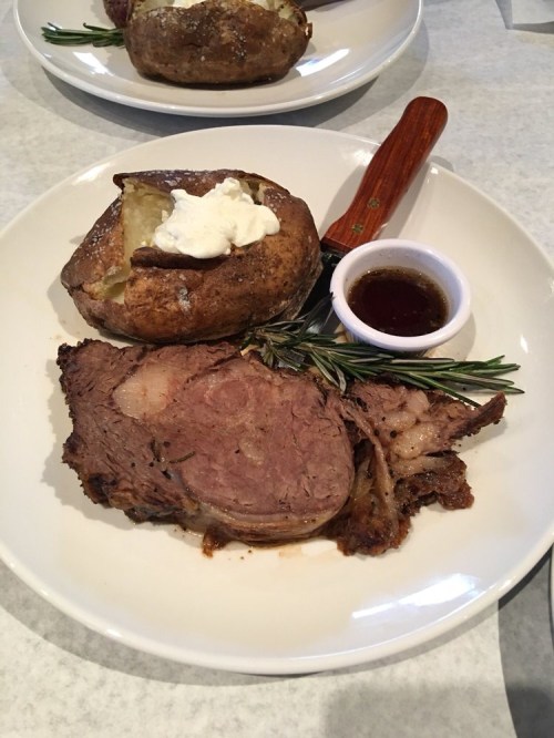 A plate featuring sliced roast beef, a baked potato with sour cream, and a small dish of sauce, garnished with rosemary.