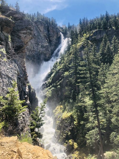 A majestic waterfall cascades down rocky cliffs, surrounded by lush green trees under a clear blue sky.