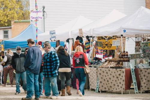 A bustling outdoor market with vendors under white tents, shoppers browsing various goods on display.