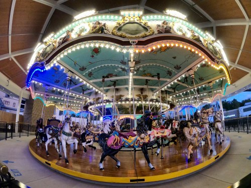A brightly lit carousel with intricately painted horses, set in a spacious area with colorful lights and decorations.