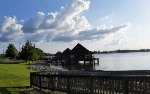A serene lakeside view featuring wooden structures, green grass, and fluffy clouds in a blue sky.