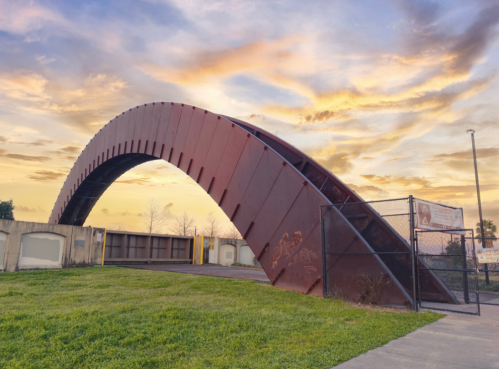 A large, curved metal bridge structure against a colorful sunset sky, with green grass in the foreground.