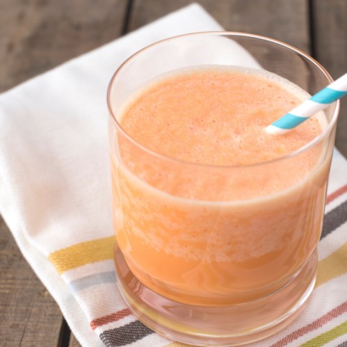 A glass of orange smoothie with a blue and white striped straw, placed on a striped napkin on a wooden table.