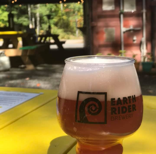 A glass of beer from Earth Rider Brewery sits on a yellow table, with a rustic outdoor setting in the background.