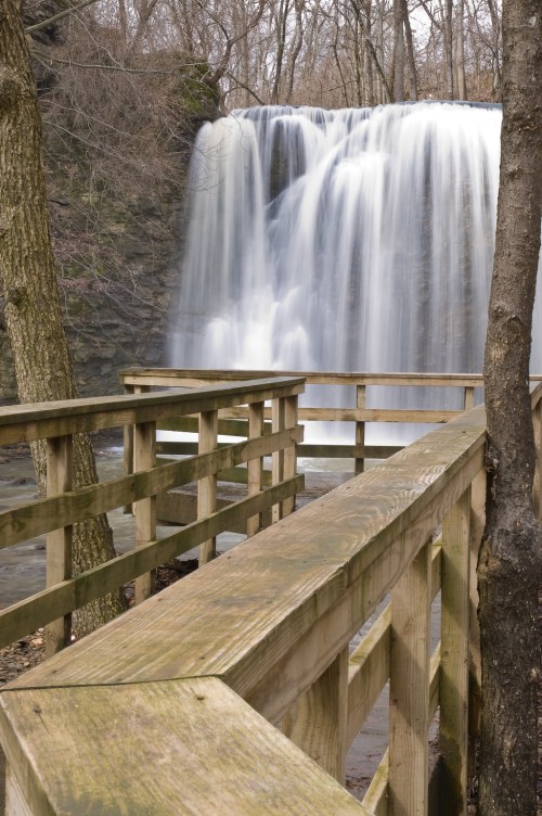 A serene waterfall cascades over rocks, surrounded by trees, with a wooden walkway in the foreground.