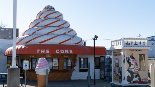 A whimsical ice cream shop with a large swirl cone roof and colorful signage, featuring a serving window and a nearby kiosk.