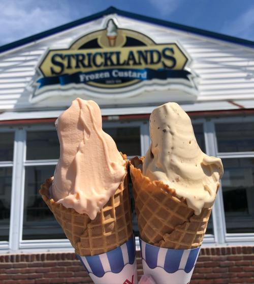 Two ice cream cones in front of a Stricklands Frozen Custard shop, one peach and one vanilla with chocolate chips.