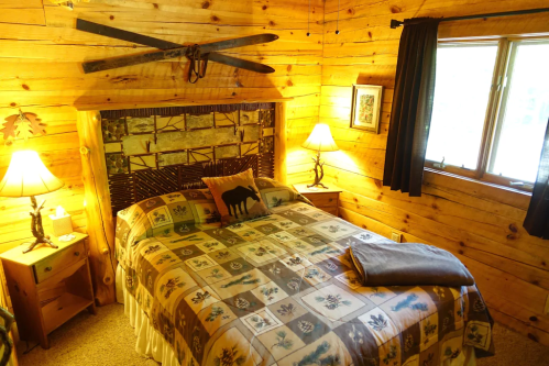 Cozy wooden bedroom with a patterned quilt, two lamps, and a window, featuring rustic decor and warm lighting.