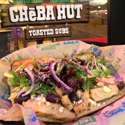 A toasted sub filled with grilled chicken, greens, olives, and onions, served in a colorful wrapper at Cheba Hut.