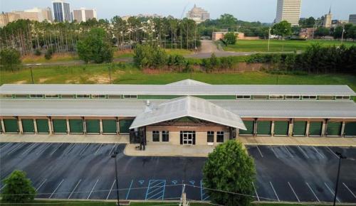 Aerial view of a storage facility with multiple units and a parking lot, surrounded by trees and city buildings in the background.