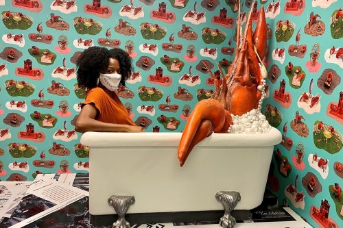 A person in an orange shirt sits in a bathtub surrounded by vibrant, quirky wallpaper and a large, artistic sculpture.
