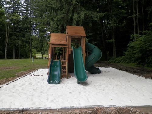 A wooden playground structure with two slides, surrounded by white sand and trees in a park setting.