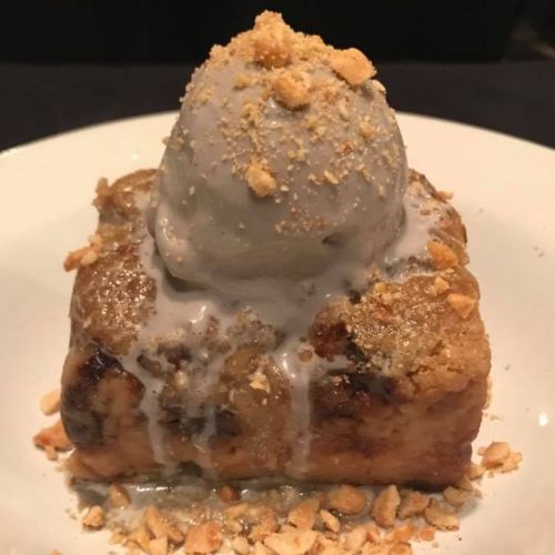 A square dessert topped with gray ice cream and crushed nuts, drizzled with a sweet sauce on a white plate.