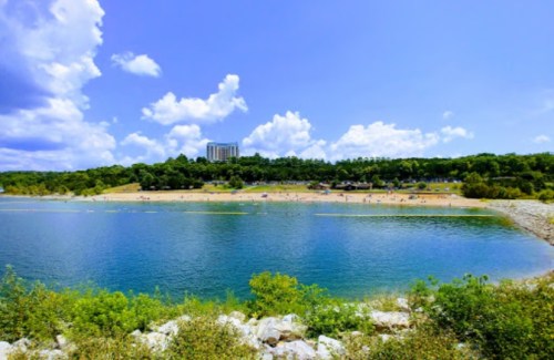 A scenic view of a beach by a lake, with people enjoying the sun and a hotel in the background under a blue sky.
