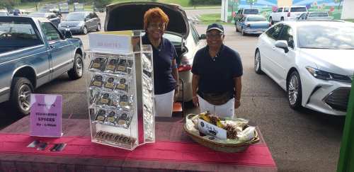 Two vendors smile behind a table displaying spice packets at an outdoor market, with cars parked nearby.