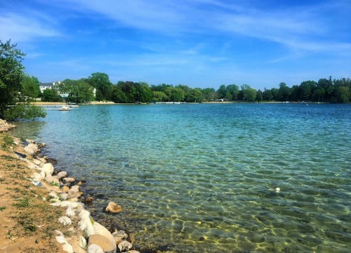 A serene lake with clear blue water, surrounded by green trees and a sandy shore under a bright blue sky.