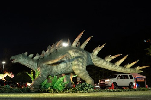 A large, illuminated dinosaur sculpture with spiked back, surrounded by greenery at night.