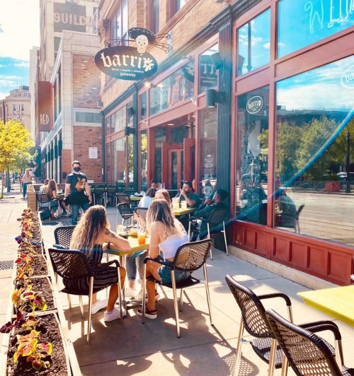 Outdoor seating at a café with people enjoying drinks, surrounded by colorful flowers and a sunny street view.