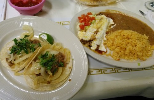 Two tacos with onions and cilantro, served with rice and a plate of beans topped with cheese and diced tomatoes.
