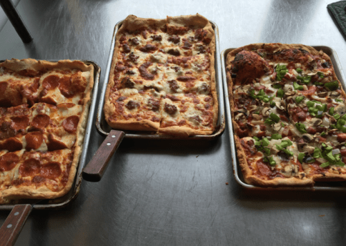 Three rectangular pizzas on a metal surface: one with pepperoni, one with sausage, and one with green peppers and mushrooms.