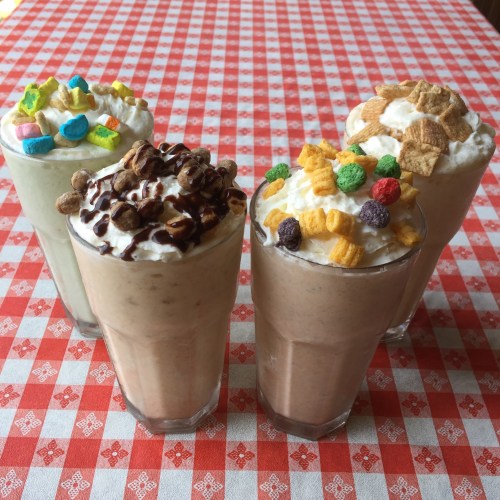 Four colorful milkshakes topped with whipped cream and various cereals, set on a red and white checkered tablecloth.
