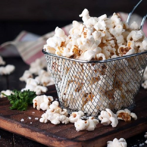 A wire basket filled with fluffy popcorn sits on a wooden board, surrounded by scattered popcorn and a sprig of parsley.