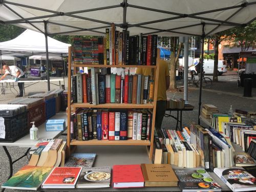 A wooden bookshelf filled with various books at an outdoor market, with tables and tents in the background.