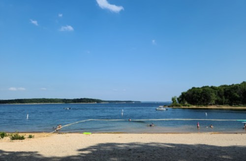 A serene lake view with a sandy beach, people swimming, and boats in the distance under a clear blue sky.