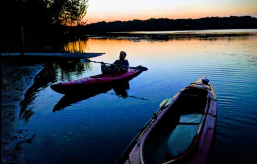 A person sits in a kayak on a calm lake at sunset, with another kayak nearby and trees silhouetted in the background.