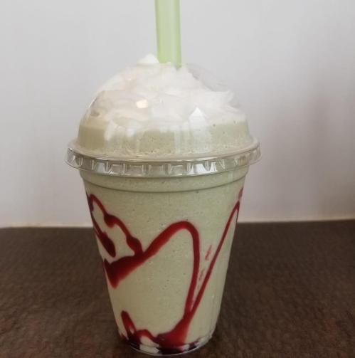 A creamy green smoothie topped with whipped cream and red syrup, served in a clear cup with a green straw.