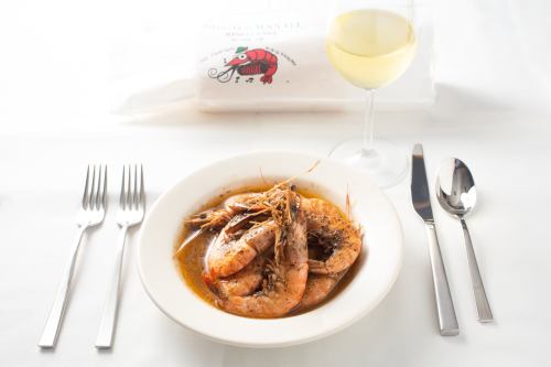 A plate of shrimp in sauce, accompanied by a glass of white wine, set on a white tablecloth with silverware.