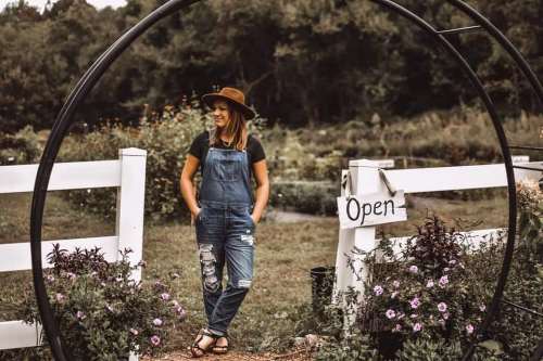 A woman in overalls and a hat stands by a garden gate with an "Open" sign, surrounded by flowers.