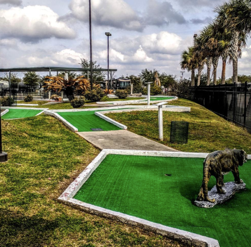 A mini-golf course with green turf, winding paths, and a tiger statue, surrounded by palm trees and cloudy skies.