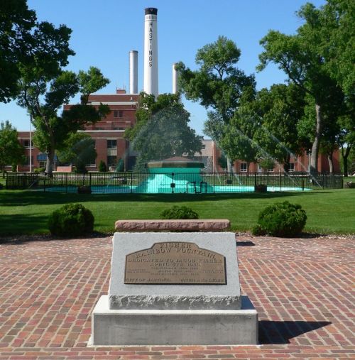 A stone monument in a park with a fountain and a factory in the background, surrounded by trees and green grass.