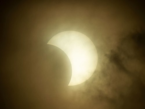 A partially eclipsed sun partially obscured by clouds, creating a dramatic silhouette against a hazy sky.