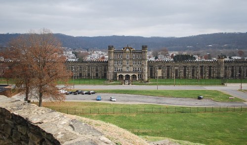 Historic building with a castle-like facade, surrounded by green grass and a parking area, under a cloudy sky.