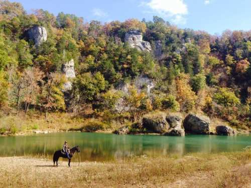 A rider on a horse stands by a tranquil lake, surrounded by colorful autumn foliage and rocky cliffs.