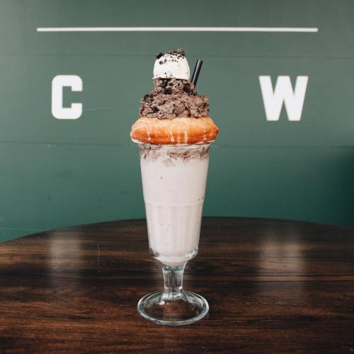 A tall glass filled with a creamy milkshake topped with a donut, whipped cream, and chocolate cookie crumbles.