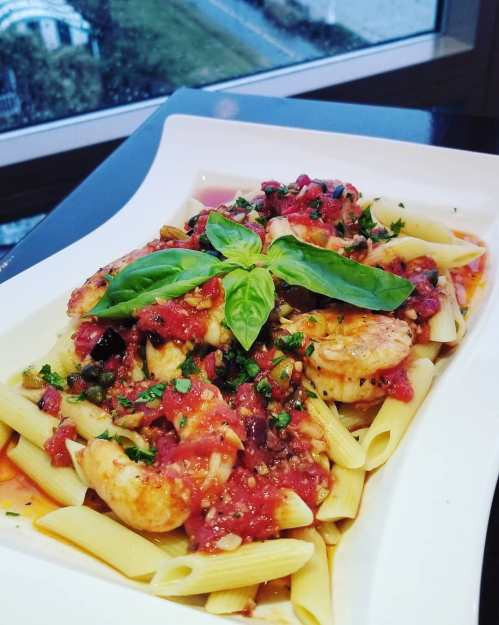 A plate of penne pasta topped with shrimp, tomato sauce, and fresh basil, set against a window view.