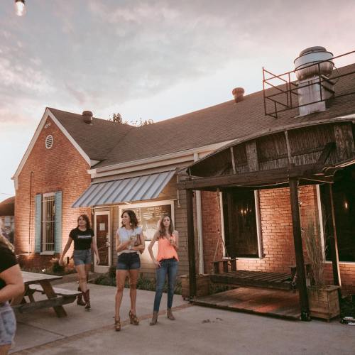 Three women walk outside a rustic brick building at sunset, enjoying a casual evening.