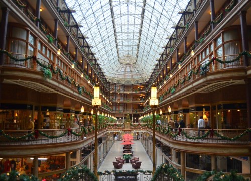 Interior view of a grand atrium with glass ceiling, decorated for the holidays, featuring multiple levels and festive greenery.