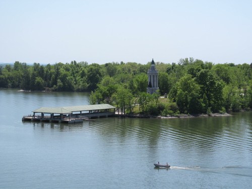 A serene lake scene with a dock, a small boat, and a monument surrounded by lush greenery under a clear blue sky.