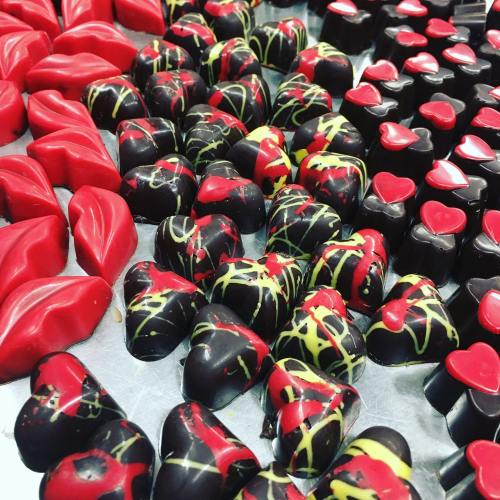 A variety of heart-shaped chocolates in red and black, some with colorful drizzles, arranged on a silver surface.