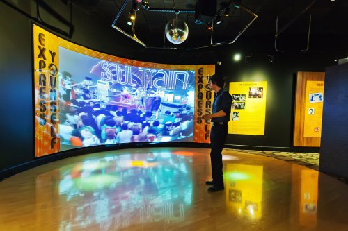A person stands in a museum exhibit with a large screen displaying a colorful music performance and a disco ball overhead.